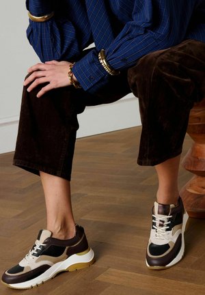 Persoon zittend op een houten kruk, draagt zwarte en bruine sneakers, donkerbruine broek, blauw gestreept overhemd en gouden armbanden.