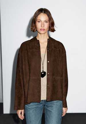 A model with short brown hair wears a brown suede shirt over a knit top, with a heart-shaped necklace, against a minimalist backdrop.