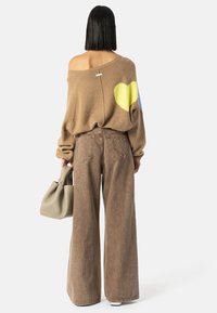 Kvinne med rett, svart hår har på seg en brun av-skulder genser med gult hjerte, vide brune bukser med hjerte på lommen, og holder en beige veske.