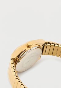 Reloj de pulsera dorado con correa de eslabones de metal, mostrando la parte trasera de la caja y la corona de cuerda sobre un fondo blanco liso.