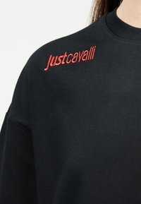 Μαύρο φούτερ με κόκκινο κεντημένο λογότυπο "Just Cavalli" στο επάνω αριστερό μέρος του στήθους κοντά στον ώμο.