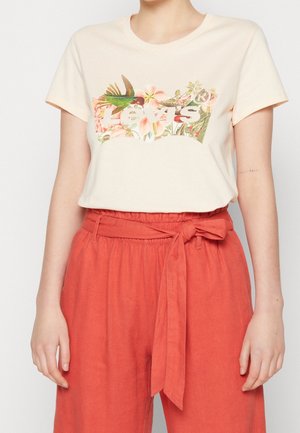 Vrouw draagt een crèmekleurig T-shirt met een bloemen Levi's-logo en een high waist oranje broek, gestrikt met een stoffen ceintuurstrik.