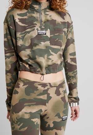 Nainen pukeutunut vihreään ja ruskeaan maastokuvioiseen lyhytlahkeiseen collegepaitaan ja yhteensopiviin legginsseihin, molemmissa Adidas-logo-patchit.