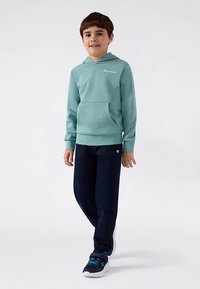 Rapaz de pé, vestindo um hoodie teal da Champion, calças de moletom azul-marinho e sapatilhas pretas com detalhes azuis, contra um fundo branco.