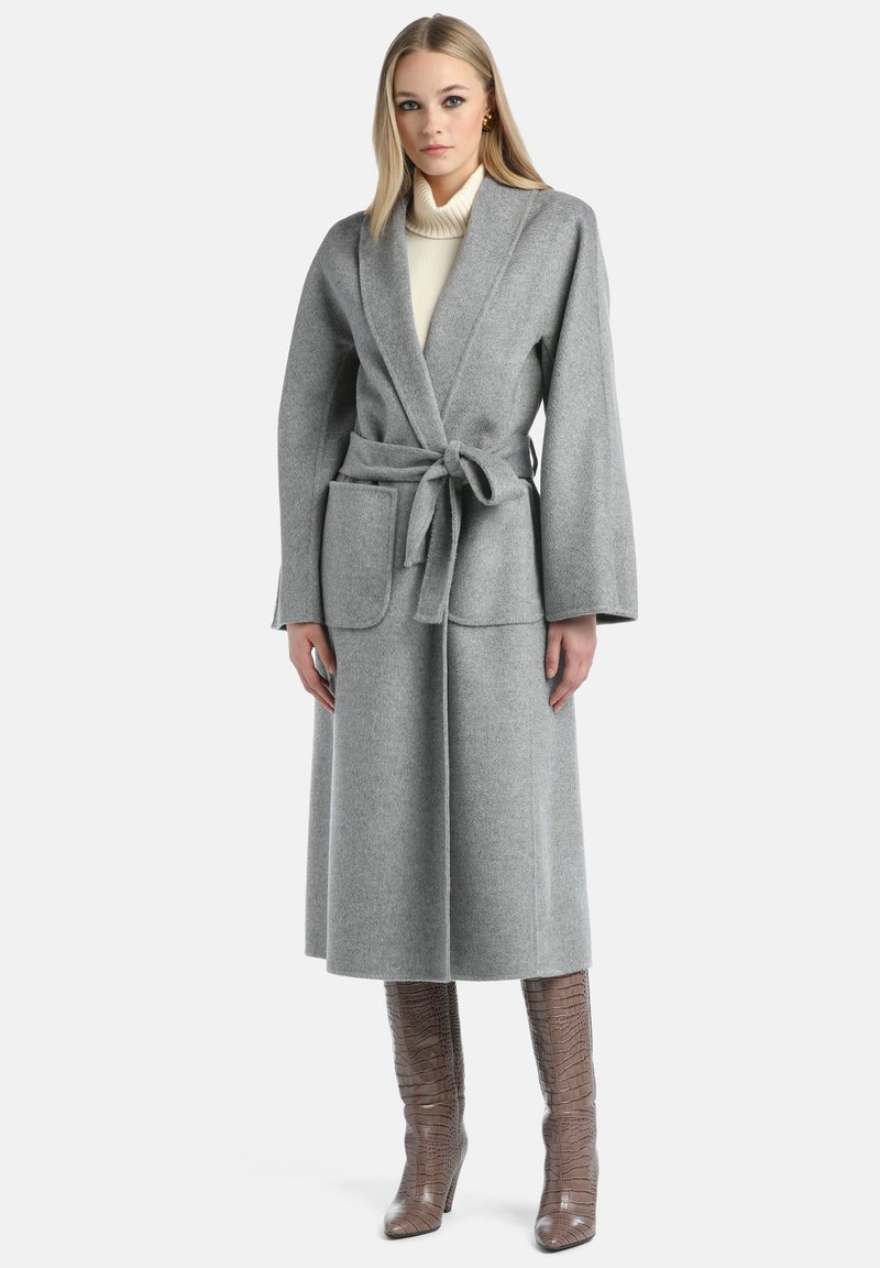 Cappotto avvolgente grigio in lana con maniche lunghe, cintura annodata, due tasche frontali e design fino al ginocchio, indossato sopra un dolcevita color crema. Linee pulite e texture.