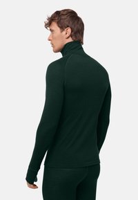 Uomo che indossa un maglione a collo alto aderente verde scuro e pantaloni abbinati, in piedi con vista di tre quarti di schiena su uno sfondo semplice.