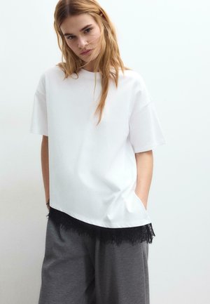 Femme aux longs cheveux blonds portant un haut blanc à manches courtes surdimensionné avec un ourlet en dentelle noire et un pantalon gris ample, debout devant un fond uni.