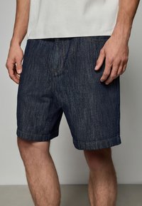 Shorts din denim de culoare albastru închis, cu un finisaj texturat, având margini rulate, buzunare tradiționale și detalii de cusătură portocalie contrastantă.
