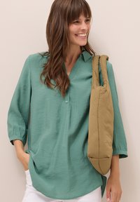 Chemisier long à manches mint avec un col à boutons et une coupe décontractée. Un sac rembourré beige repose sur l'épaule. Un pantalon blanc est visible en bas.