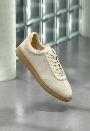 Beige mockasinsneaker med texturerad sula i gummi. Har flata snören, rundad tå och diskret logotyp på sidan. Lätt vadderad krage.
