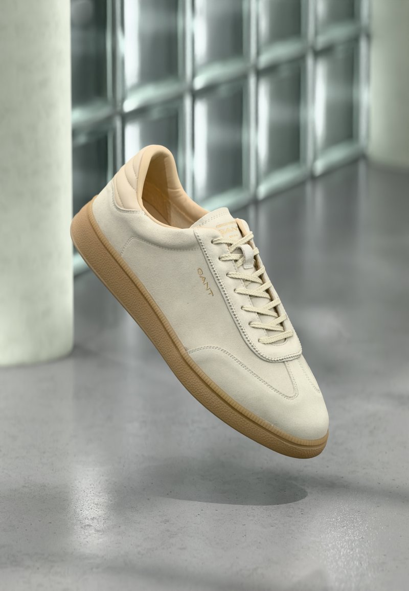 Beige suède sneaker met een gestructureerde gumrubberen zool. Heeft platte veters, een afgeronde neus en subtiele branding aan de zijkant. Licht gepolsterde kraag.