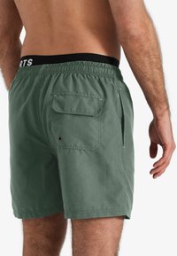 Groene zwembroek met een elastische zwarte tailleband met witte tekst. Beschikt over een achterzak en zijopeningen. Gemaakt van lichtgewicht stof.