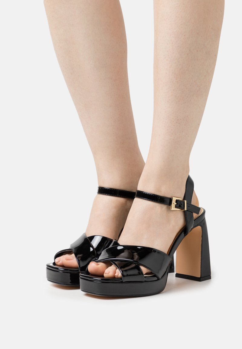 Pieds portant des talons compensés en cuir verni noir avec des brides à la cheville et des boucles dorées sur un fond blanc uni.