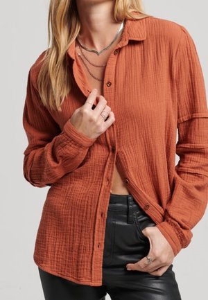 Kobieta w rdzawobrązowej teksturowanej koszuli na guziki, czarnych skórzanych spodniach, warstwowych srebrnych naszyjnikach i pierścionkach, poprawiająca kołnierzyk koszuli.