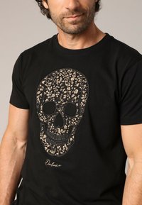 T-shirt en coton noir avec un grand graphique de crâne agrémenté de motifs floraux beiges. Comprend le mot "Deluxe" en écriture scriptée sous le design.