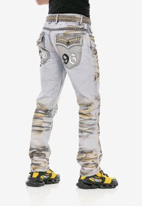 Verblasste graue Denim-Jeans mit zerstörter Textur, Metallakzenten und dekorativer Stickerei. Verfügt über Gesäßtaschen mit Verzierungen und Nummerierung.