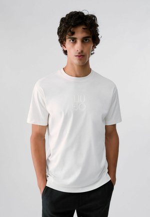 Jeune homme aux cheveux bouclés et foncés, portant un t-shirt blanc uni à manches courtes avec un texte subtil et un pantalon noir, debout sur un fond gris clair.