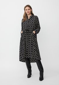 Schwarzes Kleid mit cremefarbenen Punktmuster, mit Kragen, langen Ärmeln, tailliertem Gürtel und Taschen. Getragen mit knielangen schwarzen Stiefeln.