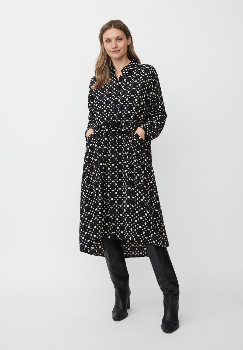 Schwarzes Kleid mit cremefarbenen Punktmuster, mit Kragen, langen Ärmeln, tailliertem Gürtel und Taschen. Getragen mit knielangen schwarzen Stiefeln.