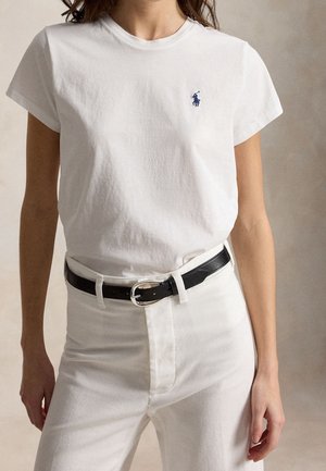 Kvinde iført en tætsiddende hvid T-shirt med korte ærmer med et lille blåt logo og hvide bukser sikret med et sort læderbælte.