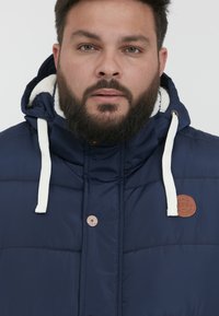 Marineblå pufferjakke med hvidt faux fur-for, tofarvede snore og et brunt læderlogo. Funktioner knaplukning.