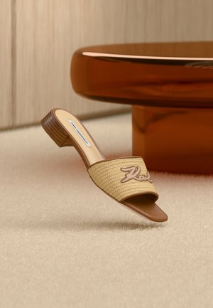 Sandalo beige intrecciato con punta aperta e tacco basso di legno, con "Karl" ricamato sulla fascia, posizionato vicino a un tavolo rotondo marrone lucido.