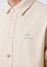 Cárdigan de punto en crema con un patrón de espiga texturizado, con un cuello puntiagudo y botones metálicos plateados. Logotipo bordado en gris.