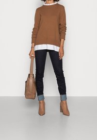 Brązowy sweter z dzianiny na białej bluzce, w połączeniu z ciemnymi dżinsami i beżowymi botkami na obcasie. Torebka na ramię w jasnobrązowym zamszowym materiale.
