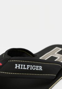 Tongs noires avec une bride texturée, arborant le logo "HILFIGER" en blanc. Semelle en caoutchouc et coutures accentuées en jaune.
