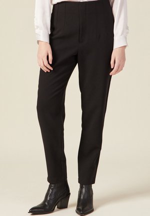 Zwarte op maat gemaakte broek met een slanke pasvorm, voorzien van plooien en een gladde textuur. Gecombineerd met zwarte enkellaarsjes met hak.