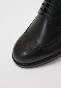 Juodas odinis formalus batų modelis su sparnų forma, turintis perforuotus raštus ir lygų paviršių. Apvalus pirštas ir tvirtas padas.