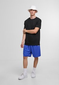 Sort bomulds t-shirt, blå athletic shorts med logo, hvid bucket hat, hvide sneakers, stående med armene korslagt mod en ensfarvet baggrund.
