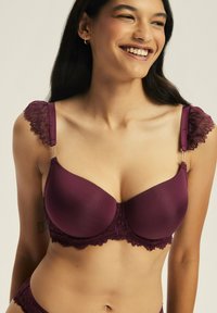 Burgundowy biustonosz z gładkiego materiału i koronkowym wykończeniem, z falbaniastymi krawędziami ramiączek oraz zebranym środkiem zapewniającym wsparcie i kształt.
