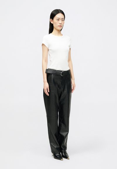 Γυναίκα που στέκεται μπροστά από λευκό φόντο, φορώντας λευκή μπλούζα με κοντά μανίκια, μαύρα παντελόνια από δέρμα με ψηλή μέση και ζώνη, και μαύρες γόβες με μυτερή μύτη.