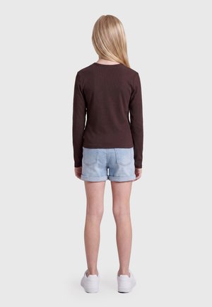 Barn med langt blondt hår iført en brun langærmet trøje, lyse denimshorts og hvide sneakers, står med ryggen mod på en hvid baggrund.