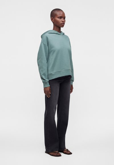 Personne debout et de face, portant un sweat à capuche vert clair, un jean foncé et des sandales marron sur un fond blanc uni.