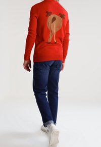 Personne portant un pull rouge avec un motif de dinde rôtie dans le dos, un jean bleu foncé et des baskets blanches, marchant de dos sur un fond uni.