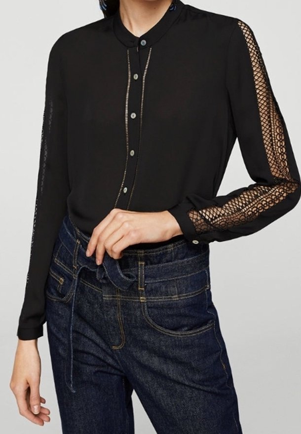 Femme portant un chemisier noir boutonné avec des manches en dentelle résille et un jean bleu foncé taille haute noué à la taille.