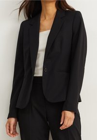 Veste blazer noire sur mesure avec un bouton unique, des revers crantés et des lignes épurées. Confectionnée dans un tissu lisse avec une silhouette ajustée.