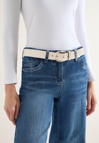 Personne portant un jean en denim bleu avec une ceinture texturée beige clair et un haut blanc à manches longues et ajusté, les mains détendues le long du corps.