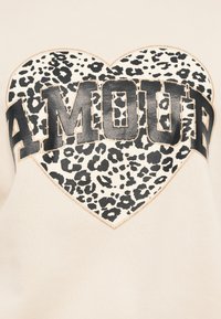 Ανοιχτό μπεζ φούτερ με μαύρο σχέδιο καρδιάς με λεοπάρδαλη και τη λέξη "AMOUR" με έντονα μαύρα γράμματα στο κέντρο.