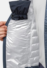 Isolierte Jacke mit glänzender silberner Futterung und einer marineblauen Tasche, ausgestattet mit einer Reißverschlusstasche und einem strukturierten blauen Außenstoff.