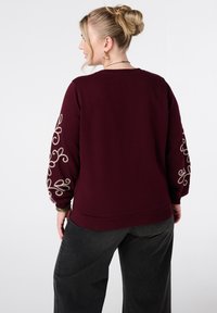 Bordeaux sweatshirt met geborduurde spiralen op de mouwen, geribbelde manchetten en zoom. Gekozen in combinatie met donkere, loszittende broeken.