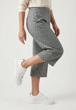 Person trägt gemusterte kurze Hose und weiße Sneaker, hebt ein im Knie gebeugtes Bein vor hellem Hintergrund.