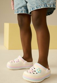 Bambino che indossa zoccoli a righe pastello con decorazioni floreali e pantaloni di jeans, in piedi su una superficie liscia con uno sfondo beige.