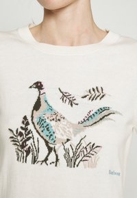 Pull blanc avec un motif brodé multicolore de faisan et de feuillage sur la poitrine, porté par une personne.