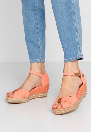 Sandalias de cuña coral con parte superior de tela anudada, correa al tobillo con hebilla y plataforma envuelta en yute. Diseño de punta cerrada que mejora la estabilidad.