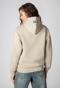 Lichtbeige hoodie met een grote capuchon en geribbelde onderrand. Heeft een merkembleem aan de achterkant van de halslijn en een gestructureerde stof. Gecombineerd met denim jeans.