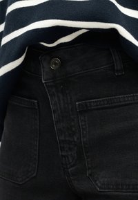 Κοντινή λήψη των πίσω τσεπών μαύρου τζιν και κουμπιού, σε συνδυασμό με ένα μπλε ναυτικό και λευκό ριγέ μπλουζάκι.