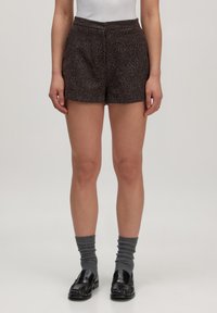 Vrouw in donkerbruin gestructureerde shorts, grijze opgerolde sokken en zwarte leren loafers die tegen een effen achtergrond staat.